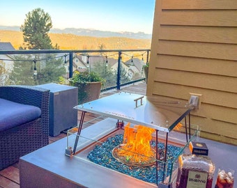 May include: A stainless steel fire pit with a glass top and a blue glass fire pit insert. The fire pit is on a deck with a view of a forest and mountains. There is a bottle of Disaronno liqueur and a glass of dark liquid on the table next to the fire pit.