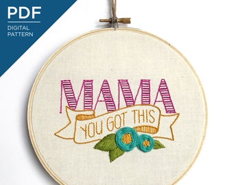 May include: Embroidered hoop art with the word "MAMA" in pink and purple with a banner underneath that reads "YOU GOT THIS" in yellow. The banner is decorated with two teal flowers and green leaves.