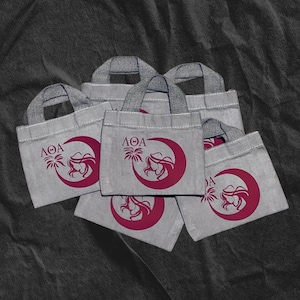 Puede incluir: Seis bolsas de tela grises con un logotipo rosa que representa una luna creciente, una palmera y el perfil de una mujer. El logotipo también incluye el texto "ΛΟΔΑ".