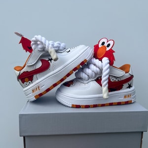 May include: White Nike Air Force 1 sneakers with red and orange accents, featuring a cartoon character design. The shoes have white laces and are displayed on a gray box. The Nike logo is visible.
