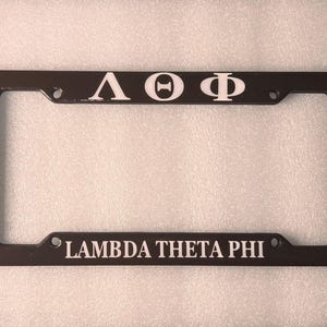 May include: A brown metal licence plate frame with white lettering that reads "Lambda Theta Phi" and "&Lambda;&Theta;&Phi;".