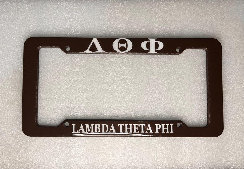 May include: A brown metal licence plate frame with white lettering. The letters "&Lambda;&Theta;&Phi;" are on the top and "Lambda Theta Phi" are on the bottom.