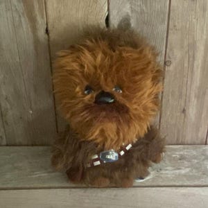 Op de afbeelding: Een pluche speelgoed van een Wookiee-personage, met bruine vacht, een zwarte neus en een bandolier. Het speelgoed wordt gepresenteerd tegen een houten achtergrond, die het gedetailleerde ontwerp en vakmanschap benadrukt.
