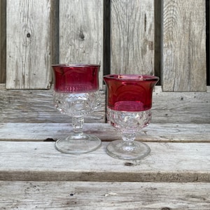 May include: Two clear glass stemmed drinking glasses with a ruby red coloured base. The glasses have a diamond pattern on the base and a rounded top.