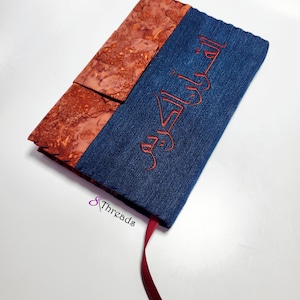 Puede incluir: Una funda de libro hecha a mano con un panel azul denim bordado con escritura árabe roja. La funda presenta un panel texturizado de color marrón rojizo y un marcapáginas de cinta roja a juego. El logotipo "8 Threads" es visible.
