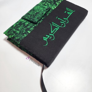 Puede incluir: Funda de libro de tela negra y verde con escritura árabe verde bordada. La solapa superior y el forro interior presentan un estampado floral verde. Una cinta negra de marcador se extiende desde la parte inferior. La marca "8 Threads" está impresa en morado.