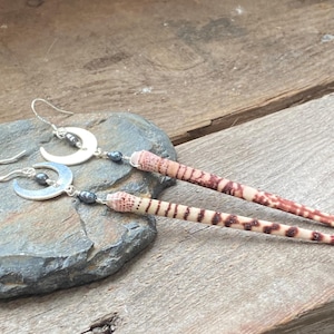 May include: A pair of dangle earrings featuring silver-toned crescent moon charms, small dark gray beads, and long, slender, tapered sea urchin spines with a natural tan and reddish-brown spotted pattern, suspended from silver-toned ear wires.