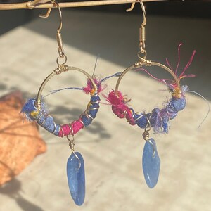 May include: A pair of handmade hoop earrings. The gold-toned hoops are wrapped with blue and pink fabric and adorned with blue gemstone pendants. The earrings hang from gold-colored ear wires, creating a unique and colorful accessory.