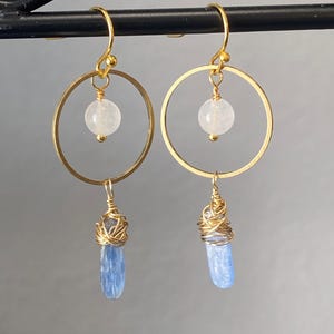 May include: A pair of gold-colored hoop earrings. Each earring features a small, round, white gemstone suspended within the hoop. Below, a blue, elongated crystal is wrapped in gold wire, creating a layered effect. The earrings hang from gold-colored hooks.