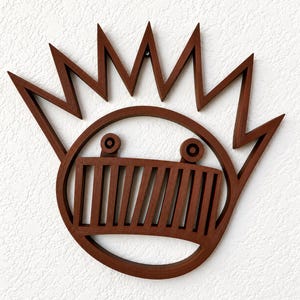 May include: A brown wooden wall art piece featuring a circular frame with a jagged, starburst-like top edge. Inside the circle, a rectangular grill-like section is positioned below two small circular eyes, creating a stylized face design.