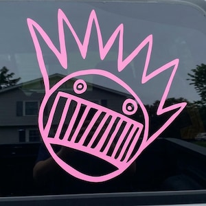 May include: A bright pink vinyl decal applied to a glass surface, featuring a stylized circular face with a large, rectangular mouth filled with vertical lines, two small circular eyes, and a jagged, spiky hair-like shape extending upward.