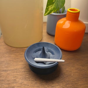 Peut inclure: Un petit cendrier en céramique bleu foncé avec une cigarette blanche à l'intérieur. Le cendrier a un centre pointu et une surface texturée.