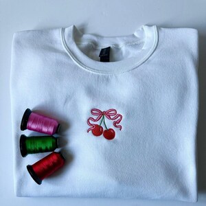 May include: A white crewneck sweatshirt featuring a small embroidered design of two red cherries tied together with a pink ribbon. Three spools of embroidery thread in pink, green, and red are positioned to the left of the cherry graphic.