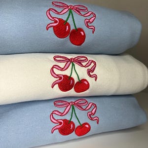 May include: A stack of three folded sweatshirts, with the top and bottom garments in light blue and the middle garment in off-white. Each sweatshirt features an identical embroidered design of two red cherries with green stems tied together with a pink ribbon.