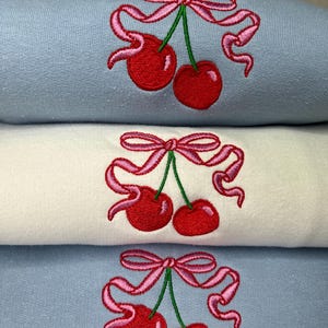 May include: A stack of three folded sweatshirts, two in light blue and one in white, each featuring an embroidered design of two red cherries with green stems tied together by a pink ribbon with flowing ends.