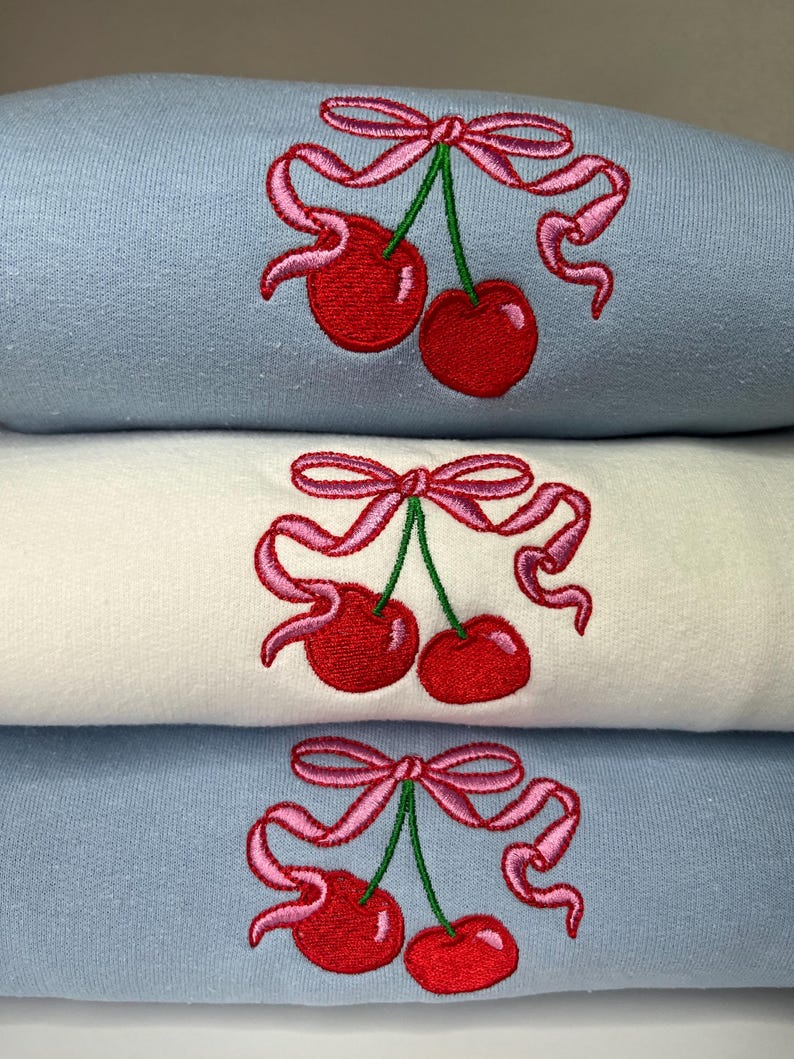 May include: A stack of three folded sweatshirts, two in light blue and one in white, each featuring an embroidered design of two red cherries with green stems tied together by a pink ribbon with flowing ends.