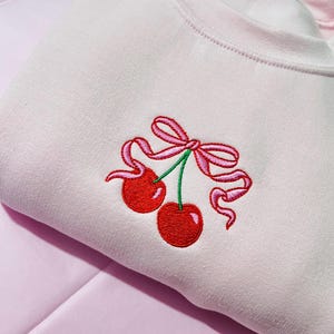May include: A light pink sweatshirt featuring an embroidered design of two bright red cherries with green stems, tied together with a decorative pink ribbon bow. The embroidery is centered on the chest area of the garment.