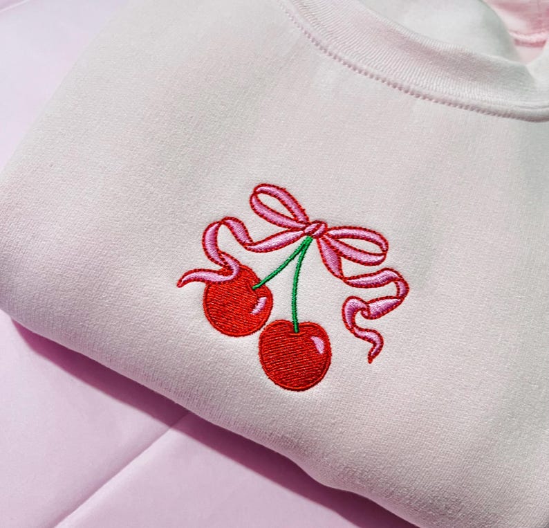 May include: A light pink sweatshirt featuring an embroidered design of two bright red cherries with green stems, tied together with a decorative pink ribbon bow. The embroidery is centered on the chest area of the garment.