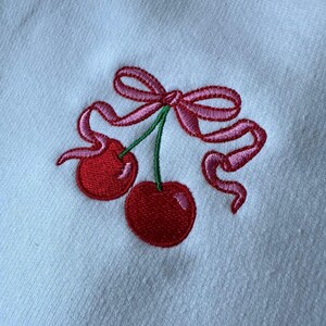 May include: A white fabric surface featuring an embroidered design of two bright red cherries with green stems. The cherries are tied together with a decorative pink ribbon that forms a bow above the fruit, with long, flowing ribbon ends.