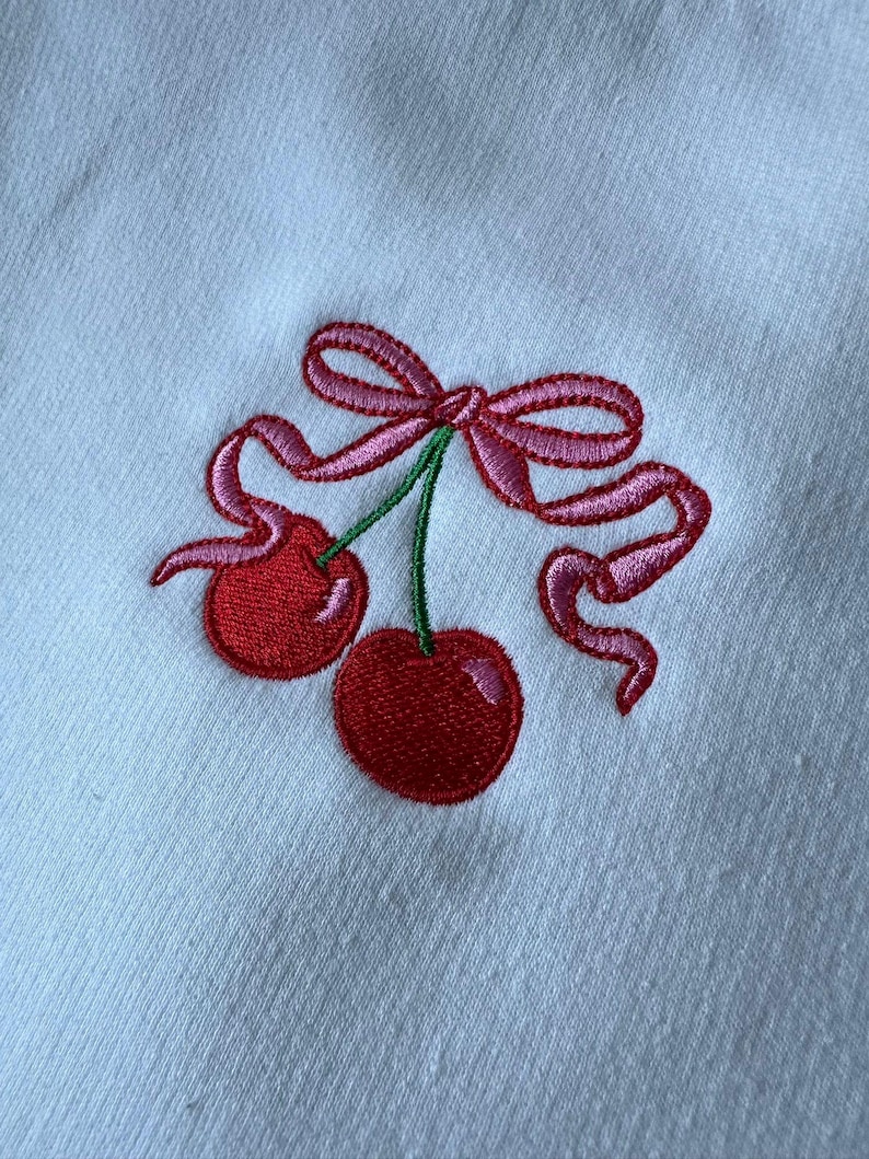May include: A white fabric surface featuring an embroidered design of two bright red cherries with green stems. The cherries are tied together with a decorative pink ribbon that forms a bow above the fruit, with long, flowing ribbon ends.