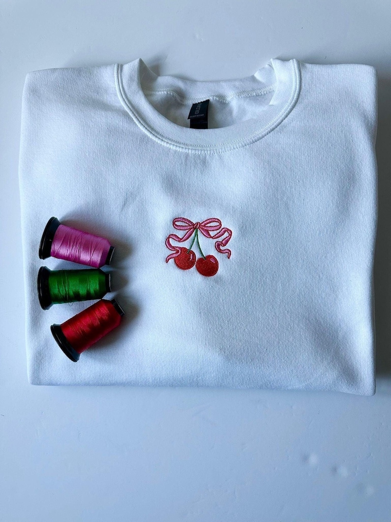 May include: A white crewneck sweatshirt featuring a small embroidered design of two red cherries tied together with a pink ribbon. Three spools of embroidery thread in pink, green, and red are positioned to the left of the cherry graphic.