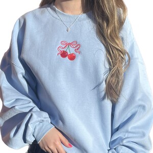 May include: A light blue crewneck sweatshirt featuring an embroidered design of two red cherries tied together with a pink ribbon bow centered on the chest. The garment has long sleeves and a ribbed collar, providing a simple and clean aesthetic.