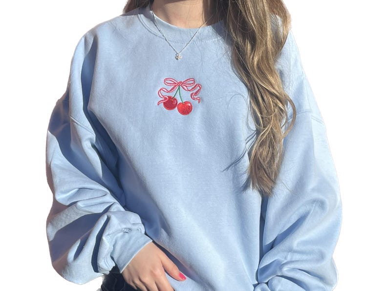 May include: A light blue crewneck sweatshirt featuring an embroidered design of two red cherries tied together with a pink ribbon bow centered on the chest. The garment has long sleeves and a ribbed collar, providing a simple and clean aesthetic.
