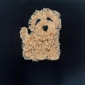 May include: A tan, textured, embroidered dog patch with black eyes and nose, set against a black background. The dog is depicted in a friendly pose, with one paw raised. The patch is likely for clothing or accessories.