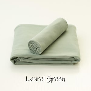 May include: A folded laurel green fabric blanket with a smooth, matte texture. A smaller, tightly rolled section of the same laurel green fabric rests diagonally on top of the folded base. The text Laurel Green is printed below the fabric.