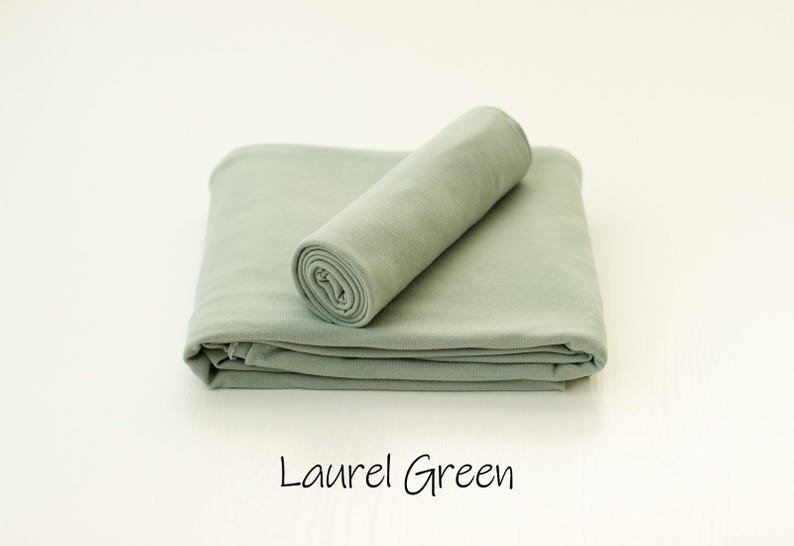 May include: A folded laurel green fabric blanket with a smooth, matte texture. A smaller, tightly rolled section of the same laurel green fabric rests diagonally on top of the folded base. The text Laurel Green is printed below the fabric.