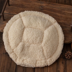 May include: A round, cream-colored, fluffy cushion with a quilted design. The cushion is made of soft, plush fabric and has a slightly raised center.