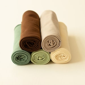 May include: A collection of five fabric rolls arranged in a cluster. The textiles feature a palette of muted earth tones, including sage green, dark chocolate brown, beige, light olive green, and a soft cream color, all tightly rolled into cylinders.