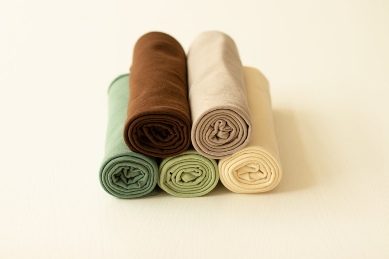 May include: A collection of five fabric rolls arranged in a cluster. The textiles feature a palette of muted earth tones, including sage green, dark chocolate brown, beige, light olive green, and a soft cream color, all tightly rolled into cylinders.