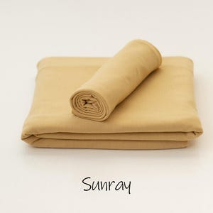 May include: A solid light tan fabric piece folded into a rectangular shape with a smaller matching fabric roll placed on top. The word Sunray is printed in black text below the fabric, indicating the specific color of the textile material.