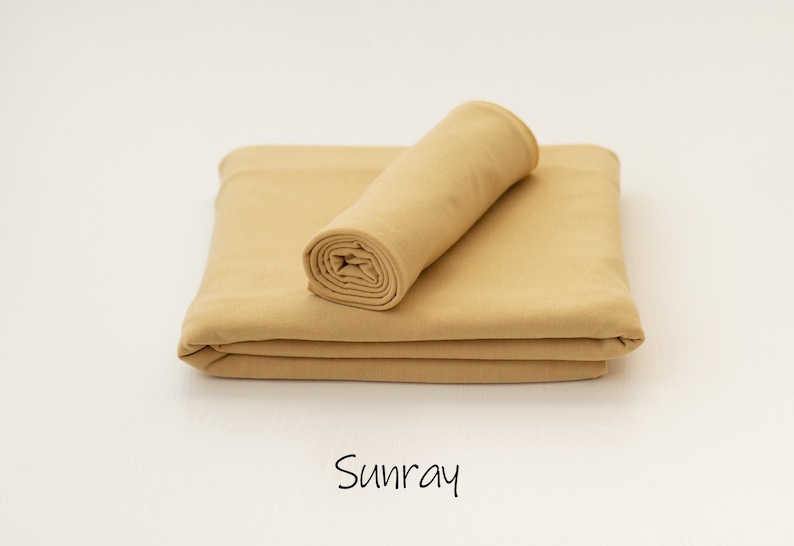 May include: A solid light tan fabric piece folded into a rectangular shape with a smaller matching fabric roll placed on top. The word Sunray is printed in black text below the fabric, indicating the specific color of the textile material.
