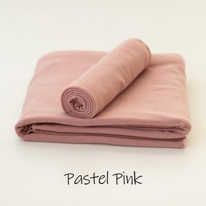 May include: A folded piece of solid pastel pink fabric with a matching rolled fabric cylinder placed on top. The text "Pastel Pink" is printed in black below the fabric, which features a smooth, matte texture and consistent color throughout.