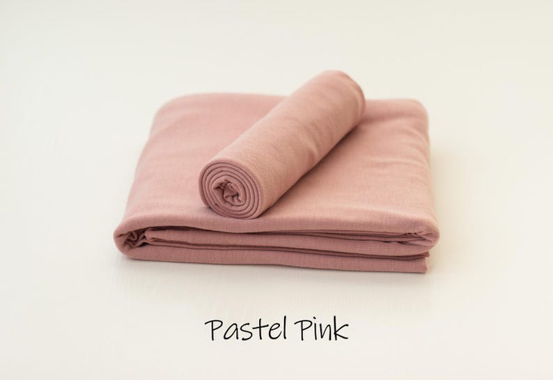 May include: A folded piece of solid pastel pink fabric with a matching rolled fabric cylinder placed on top. The text "Pastel Pink" is printed in black below the fabric, which features a smooth, matte texture and consistent color throughout.
