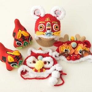 May include: Five red, white and yellow crocheted lion dance hats with embroidered details. The hats are on a white surface.