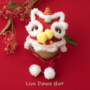 May include: A red and white crocheted lion dance hat with yellow and blue accents. The hat has a fluffy white trim and two white pom-poms attached to the strings.  Lion Dance Hat.