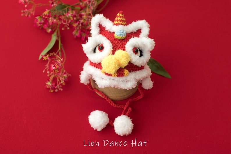 May include: A red and white crocheted lion dance hat with yellow and blue accents. The hat has a fluffy white trim and two white pom-poms attached to the strings.  Lion Dance Hat.