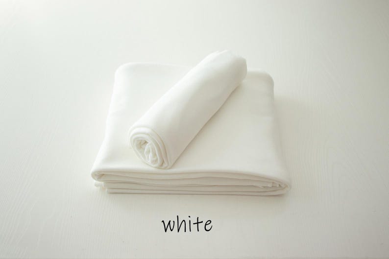May include: A solid white fabric textile folded into a stack with a single rolled piece resting on top. The word white is printed in black lowercase letters below the fabric, indicating the color of the material shown in the image.