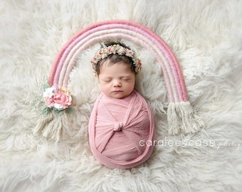 May include: A newborn baby girl is swaddled in a pink fabric and wearing a flower crown. The baby is lying on a white fluffy surface with a pink and white rainbow prop behind her.