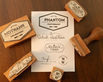 May include: Wooden stamps with various designs and text, including "PHANTOM PHOTOGRAPHY NEW YORK" and "BOTANICAL FORMS." The stamps are arranged around a sheet of paper with similar text and logos. The stamps are made of wood and have a vintage aesthetic.