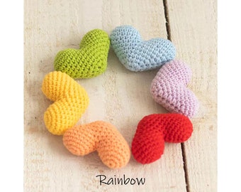 May include: A rainbow of crocheted hearts in various colors, including red, orange, yellow, green, blue, and purple. The hearts are arranged in a circle on a white wooden surface.