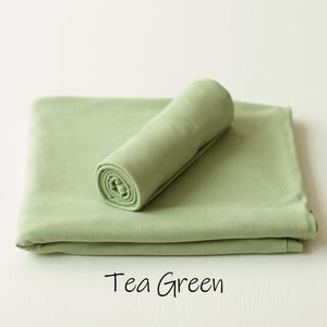 May include: A tea green fabric wrap featuring a smooth, solid-colored textile surface. The item consists of one piece folded flat into a rectangular shape with a second piece rolled into a cylinder and placed on top. The text Tea Green is visible.