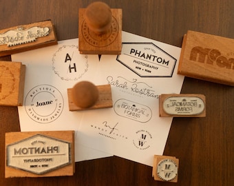 May include: A collection of wooden stamps with various designs and text, including logos and names. The stamps are arranged on a wooden surface with printed paper. The designs include the words "Phantom Photography", "Botanical Forms", and "Joane Handmade Jewelry".