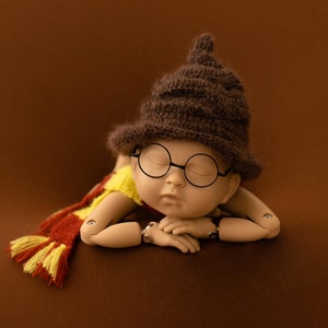 May include: A sleeping baby doll wearing a brown knitted hat with a pointed top and round black glasses. The doll is wearing a yellow and red scarf.