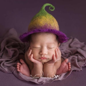 Peut inclure: Un nouveau-né portant un bonnet en laine violet et vert avec un sommet vert bouclé, est allongé sur un tissu violet. Le bébé dort les mains sous le menton.