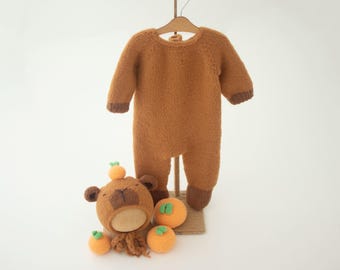 May include: A brown knitted baby onesie with long sleeves and a bear hat. The hat has a brown face and green leaves on top. There are also four small orange and green fruit decorations.