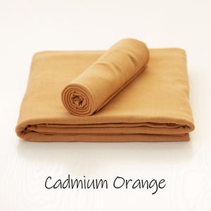 May include: A cadmium orange fabric cloth folded into a rectangular shape with a smaller matching piece rolled and placed on top. The text Cadmium Orange is printed in a simple black font below the fabric, indicating the specific color name.